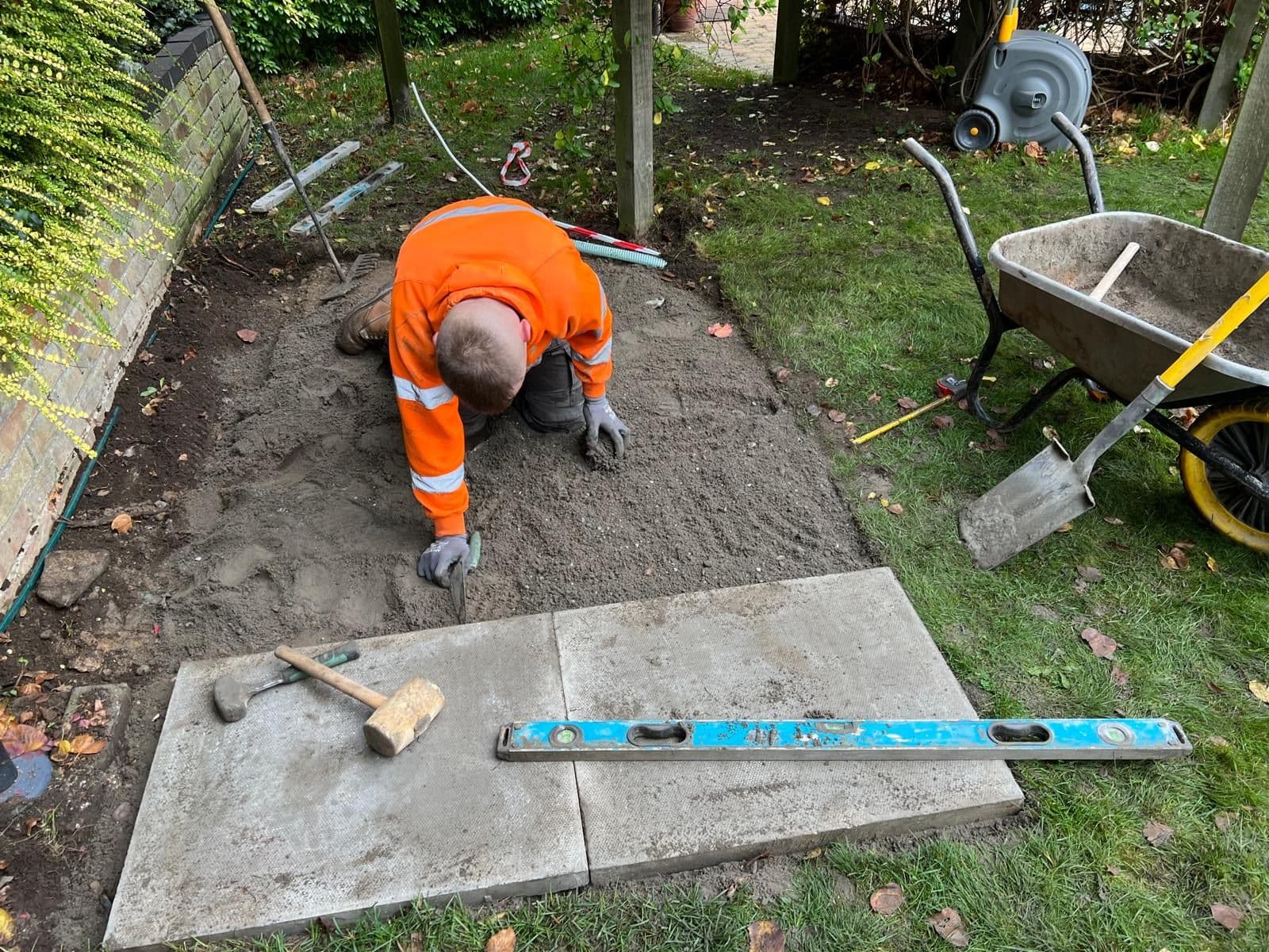 An engineer in hi-vis kneeling to level a sand bed before laying the paving slabs for a new oil tank base, with a spirit level and wheelbarrow alongside