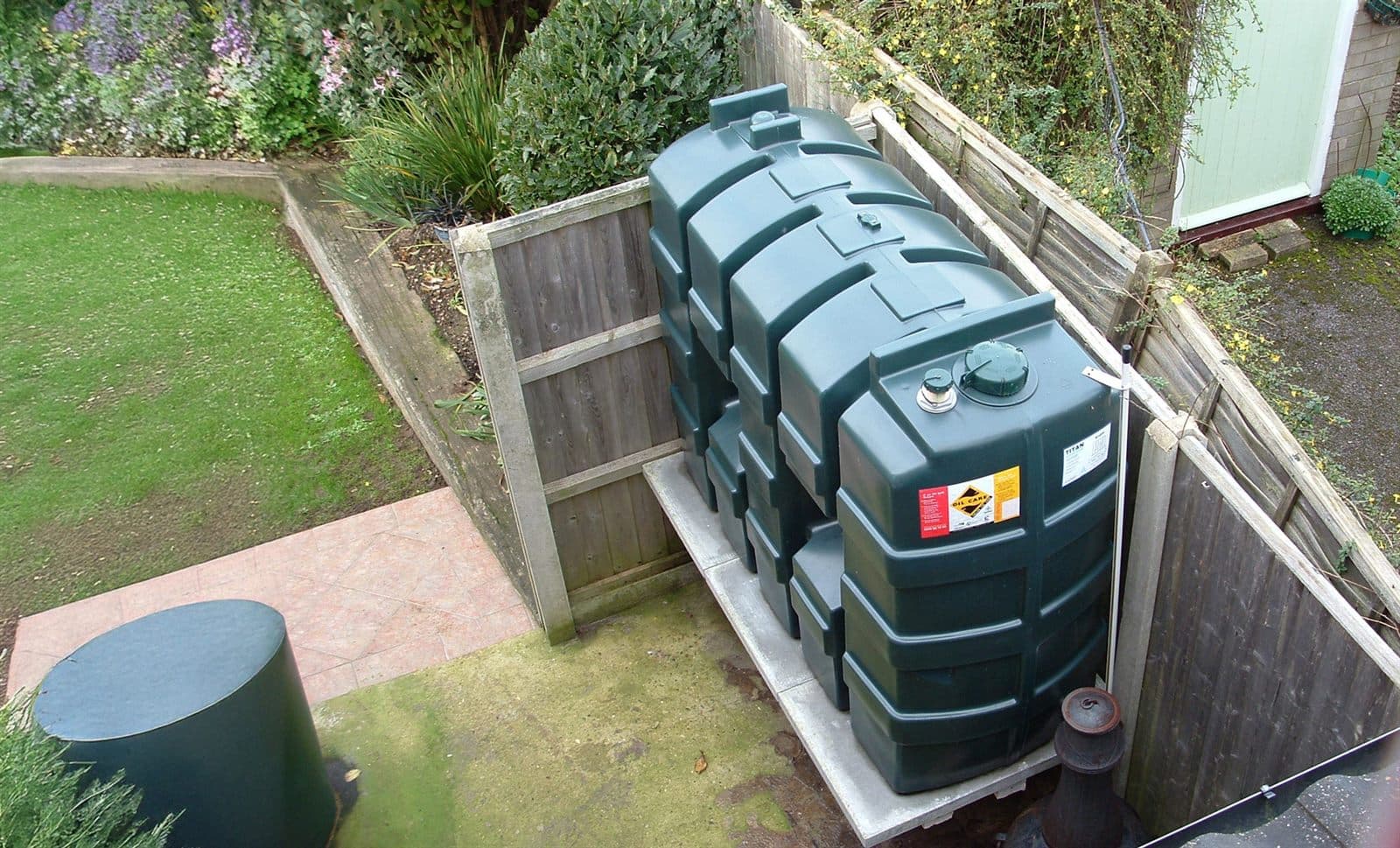 A green bunded oil tank installed on a concrete base in a fenced corner of a back garden, seen from above