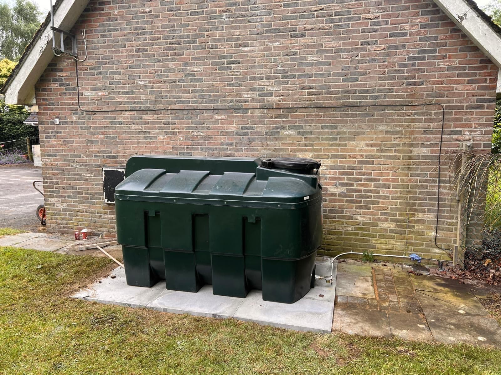 A green bunded oil tank installed on a level paving-slab base against the brick gable of a house, with the oil line run along the wall