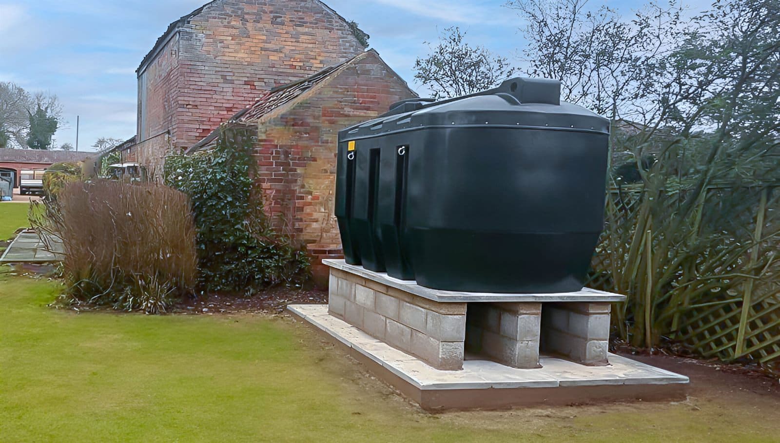 A large green bunded oil tank on a newly built blockwork and paving-slab plinth in a lawned garden, with a brick farm building behind